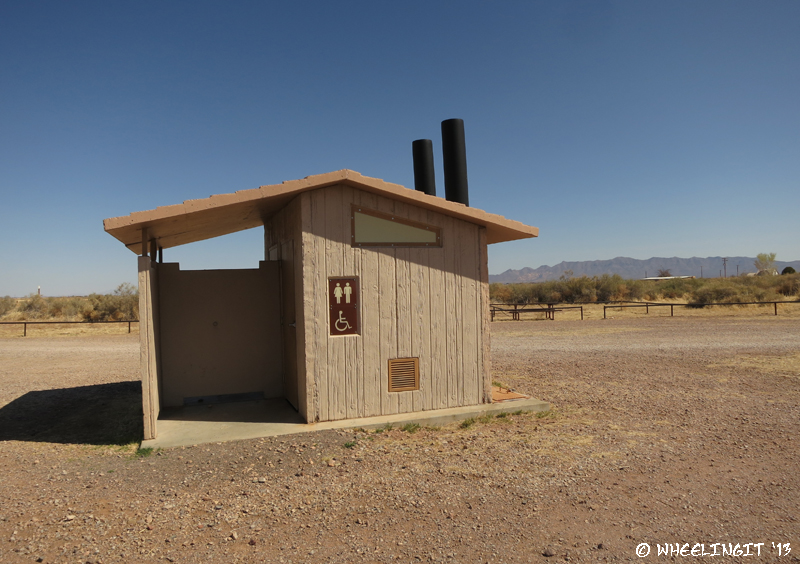 View of pit toilets in middle of camping area. There is a also a garbage disposal.